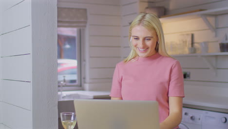 Woman-Putting-In-Wireless-Earbuds-Standing-At-Kitchen-Counter-Making-Video-Call-On-Laptop