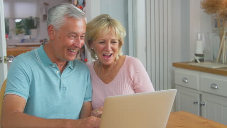 Senior-Retired-Couple-Sitting-Around-Table--At-Home-Making-Video-Call-On-Laptop