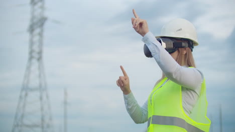 A-woman-electrician-in-virtual-reality-glasses-moves-her-hand-simulating-the-work-with-the-graphical-interface-of-a-power-plant-against-the-background-of-high-voltage-electric-transmission-lines.