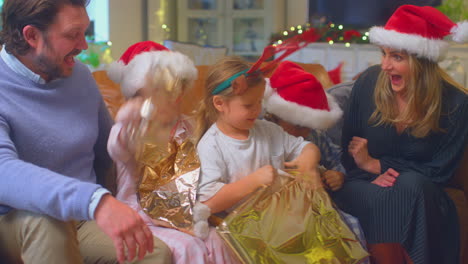 Family-Wearing-Santa-Hats-Sitting-On-Sofa-At-Home-Opening-Christmas-Gifts