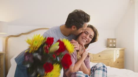Couple-Wearing-Pyjamas-In-Bed-At-Home-Celebrating-Birthday-Or-Anniversary-With-Card-And-Flowers