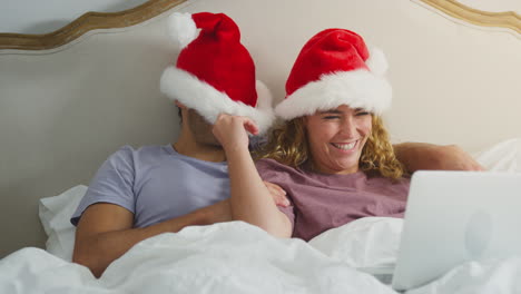 Paar-Zu-Weihnachten-Liegt-Zu-Hause-Im-Bett-Und-Führt-Videoanrufe-Auf-Laptop-Mit-Weihnachtsmützen