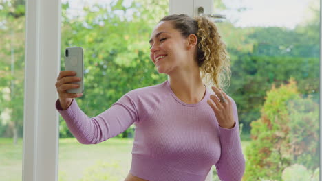Young-Woman-Sitting-On-Yoga-Mat-At-Home-After-Exercise-Posing-For-Selfie-On-Mobile-Phone