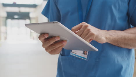 Close-Up-Of-Male-Medical-Worker-In-Scrubs-Using-Digital-Tablet-In-Busy-Hospital-Building