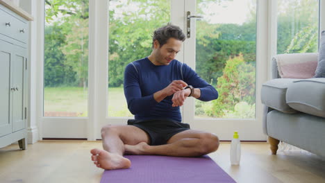 Mature-Man-Sitting-On-Yoga-Mat-At-Home-After-Exercise-Looking-At-Smart-Watch