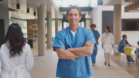 Portrait-Of-Male-Doctor-Wearing-Scrubs-Folding-Arms-And-Standing-In-Busy-Hospital