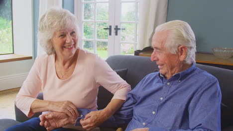Senior-Couple-Sitting-On-Sofa-At-Home-With-Woman-Helping-Man-To-Fasten-Button-On-Shirt-Cuff