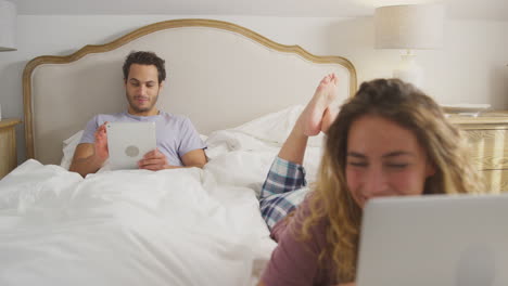 Couple-Wearing-Pyjamas-In-Bed-At-Home-With-Woman-Using-Laptop-And-Man-Digital-Tablet