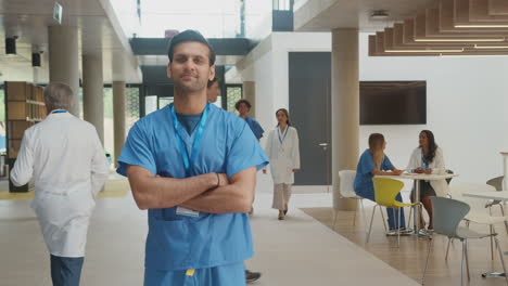 Retrato-De-Un-Médico-Masculino-Vestido-Con-Batas-Cruzando-Los-Brazos-Y-De-Pie-En-Un-Hospital-Ajetreado