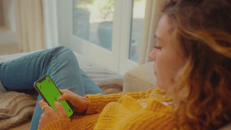 Over-The-Shoulder-View-Of-Young-Woman-At-Home-Using-Mobile-Phone-With-Green-Screen