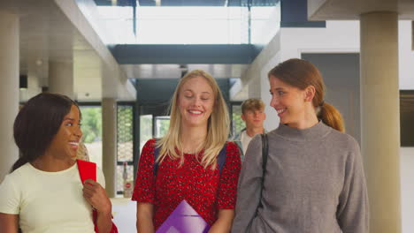 Three-Female-Students-In-Busy-University-Or-College-Building-Talking-As-They-Walk-Along-Corridor