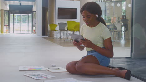 Female-University-Student-Sitting-On-Floor-Of-College-Building-Using-Digital-Tablet