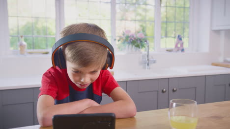 Niño-En-Casa-En-La-Cocina-Viendo-Una-Película-O-Un-Programa-En-Un-Teléfono-Móvil-Usando-Auriculares-Inalámbricos