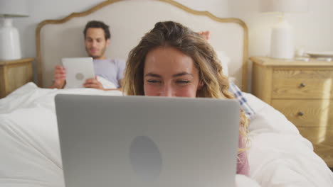 Ein-Paar-Im-Schlafanzug-Liegt-Zu-Hause-Im-Bett,-Eine-Frau-Benutzt-Einen-Laptop-Und-Ein-Mann-Ein-Digitales-Tablet