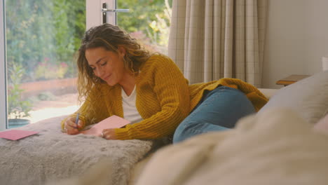 Woman-In-Cosy-Warm-Jumper-Lying-On-Sofa-At-Home-Lying-On-Sofa-Writing-Greetings-Card-Or-Letter