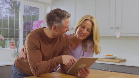 Mature-couple-at-home-in-kitchen-using-digital-tablet-to-book-show,-vacation-or-make-purchase-together---shot-in-slow-motion