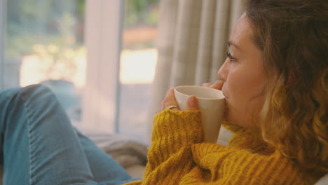 Young-Woman-In-Cosy-Warm-Jumper-Sitting-On-Sofa-At-Home-Looking-Out-Of-Window-With-Hot-Drink