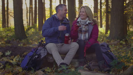 Mature-couple-sitting-on-log-stop-for-rest-and-hot-drink-on-walk-through-fall-or-winter-countryside---shot-in-slow-motion