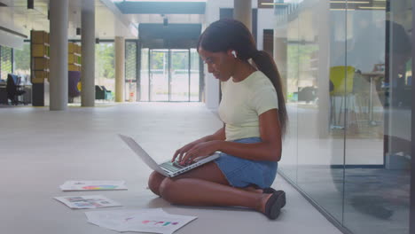 Female-University-Student-Sitting-On-Floor-Of-College-Building-Using-Laptop
