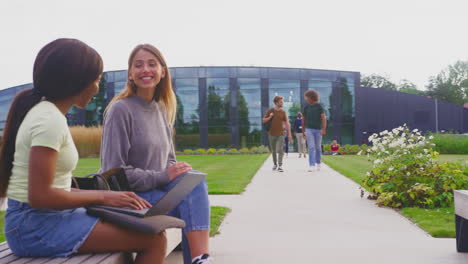 Female-University-Or-College-Students-Sitting-Outdoors-On-Campus-Talking-And-Working-On-Laptop