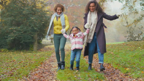 Lächelnde-Weibliche-Mehrgenerationenfamilie,-Die-Gemeinsam-Ein-Mädchen-In-Der-Herbstlichen-Landschaft-Spaziert-Und-Schaukelt