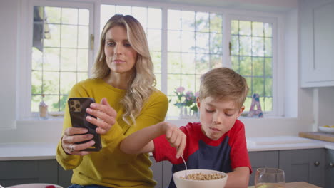 Hijo-En-Casa-Desayunando-Cereal-En-La-Cocina-Mientras-La-Madre-Hace-Una-Videollamada-Por-Teléfono-Móvil