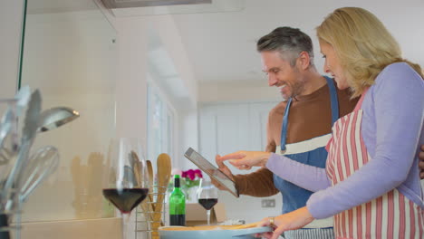 Älteres-Paar-Zu-Hause-Folgt-Einem-Rezept-Auf-Einem-Digitalen-Tablet-In-Der-Küche,-Während-Es-Gemeinsam-Eine-Mahlzeit-Zubereitet-–-Aufgenommen-In-Zeitlupe