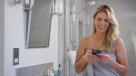 Mature-Woman-Getting-Ready-In-Bathroom-At-Home-Looking-At-Mobile-Phone-And-Laughing