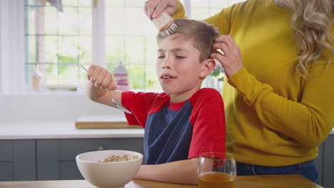 Unhappy-Son-At-Home-Eating-Breakfast-Cereal-At-Kitchen-Counter-As-Mother-Brushes-His-Hair