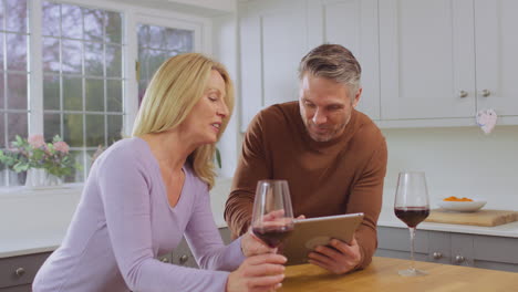 Mature-couple-at-home-in-kitchen-using-digital-tablet-to-book-show,-vacation-or-make-purchase-and-celebrating-by-drinking-red-wine-together---shot-in-slow-motion