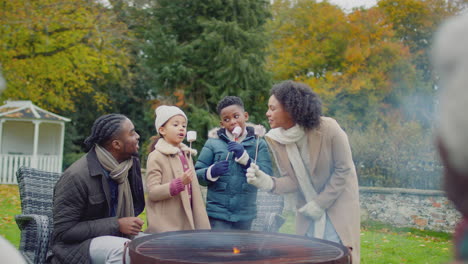 Multi-generation-family-toasting-and-eating-marshmallows-standing-around-firepit-or-barbeque-in-garden-at-home---shot-in-slow-motion