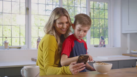 Son-At-Home-Eating-Breakfast-Cereal-At-Kitchen-Counter-Looking-At-Mobile-Phone-With-Mother