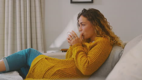 Young-Woman-In-Cosy-Warm-Jumper-Sitting-On-Sofa-At-Home-Looking-Out-Of-Window-With-Hot-Drink