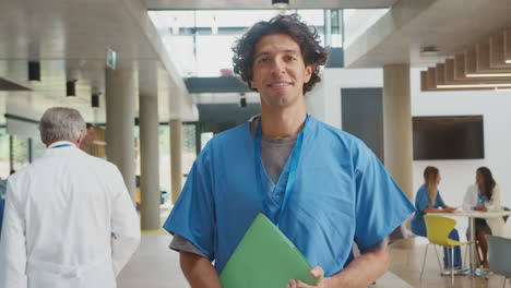 Portrait-Of-Male-Doctor-Wearing-Scrubs-Holding-Patient-Notes-In-Busy-Hospital-Building