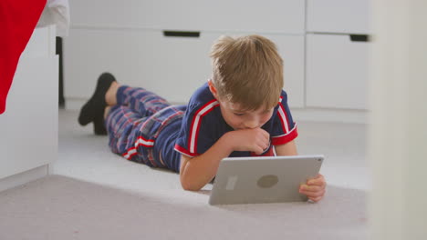 Niño-En-Casa-En-El-Dormitorio-En-Pijama-Viendo-Una-Película-O-Un-Programa-En-Una-Tableta-Digital-Tumbado-En-El-Suelo