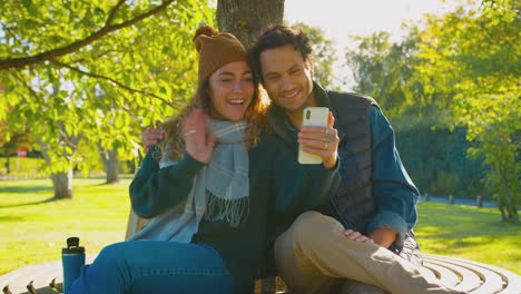 Couple-Sitting-On-Bench-In-Autumn-Park-Making-Video-Call-On-Mobile-Phone