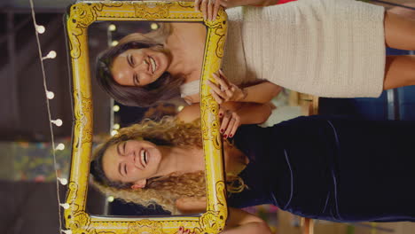 Vertical-Video-Of-Female-Friends-Having-Fun-Posing-With-Photo-Booth-Photo-Frame-At-Party-In-Bar