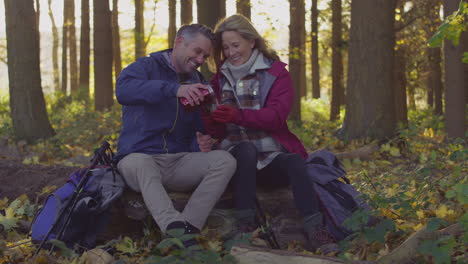 Mature-couple-sitting-on-log-stop-for-rest-and-pour-hot-drink-from-flask-on-walk-through-fall-or-winter-countryside---shot-in-slow-motion