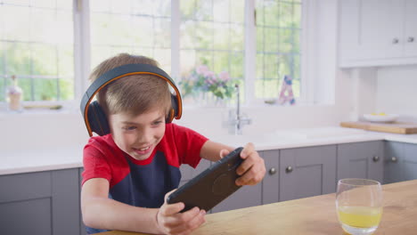 Junge-Zu-Hause-In-Der-Küche-Spielt-Ein-Spiel-Auf-Dem-Mobiltelefon-Und-Trägt-Kabellose-Kopfhörer