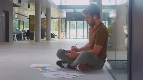 Male-University-Student-Sitting-On-Floor-Of-College-Building-Using-Digital-Tablet