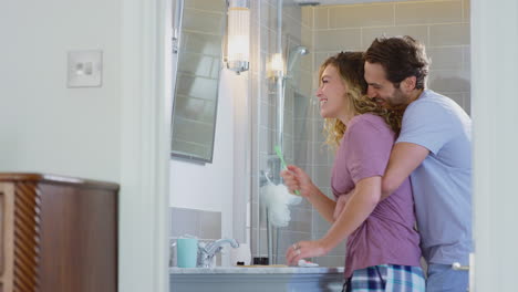 Man-Hugging-Woman-Wearing-Pyjamas-In-En-Suite-Bathroom-As-She-Brushes-Her-Teeth