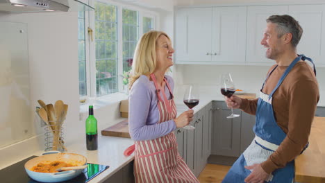Mature-couple-wearing-aprons-drinking-red-wine-as-they-prepare-ingredients-and-cook-meal-at-home-together---shot-in-slow-motion
