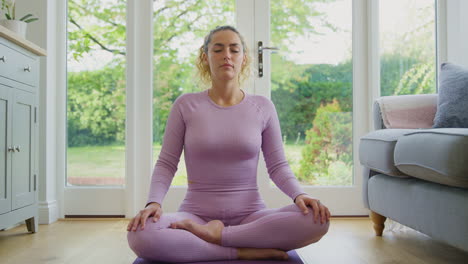 Retrato-De-Una-Joven-Sonriente-Sentada-Sobre-Una-Alfombra-En-Casa-En-Posición-De-Yoga-Meditando