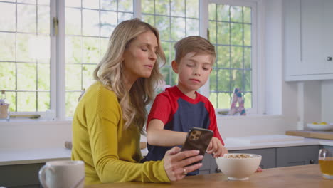 Son-At-Home-Eating-Breakfast-Cereal-At-Kitchen-Counter-Looking-At-Mobile-Phone-With-Mother