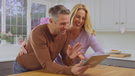Mature-couple-at-home-in-kitchen-using-digital-tablet-to-make-video-call-to-friends-or-family---shot-in-slow-motion