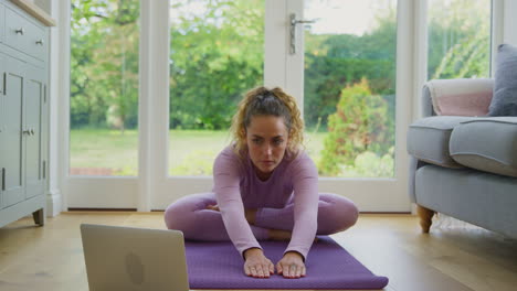 Junge-Frau-Sitzt-Zu-Hause-Auf-Einer-Matte-Mit-Laptop-Und-Macht-Einen-Online-Yoga-Kurs