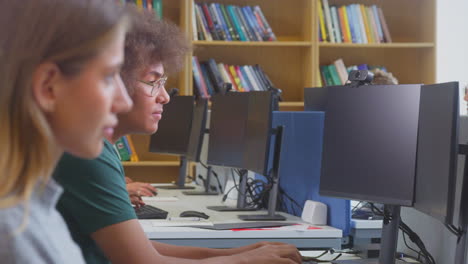 Male-University-Or-College-Student-Working-At-Computer-In-Library-Being-Helped-By-Tutor