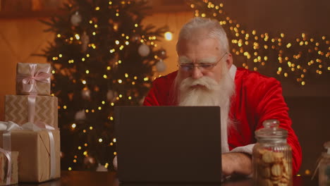 Real-Santa-Claus-using-new-technology-for-communication-with-children-receiving-mail-or-wish-list.-Cheerful-working-on-laptop-and-smiling-while-sitting-at-his-chair-with-fireplace-and-Christmas-Tree.-High-quality-4k-footage