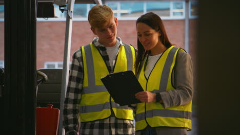 Female-Team-Leader-With-Clipboard-In-Warehouse-With-Male-Intern-Standing-By-Fork-Lift-Truck