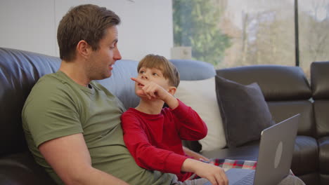 Father-and-son-sitting-on-sofa-at-home-in-pyjamas-together-looking-at-laptop---shot-in-slow-motion
